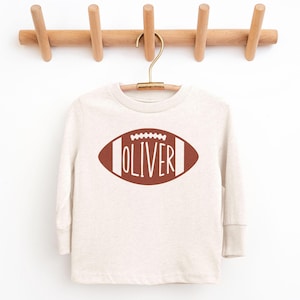 Pode incluir: Uma camisa de manga comprida creme com um gráfico de futebol americano castanho. O design da bola de futebol inclui o nome "OLIVER" em letras brancas. A camisa está pendurada em um cabide dourado em um cabideiro de madeira.