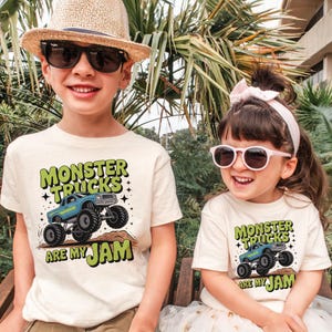 Puede incluir: Dos ni&ntilde;os con camisetas color crema con un gr&aacute;fico de monster truck. Las camisetas dicen "MONSTER TRUCKS ARE MY JAM" en verde y negro. Uno lleva gafas de sol y un sombrero de paja, el otro lleva gafas de sol y una diadema rosa.