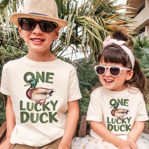 Puede incluir: Dos ni&ntilde;os con camisetas color crema con un gr&aacute;fico de pato y el texto "ONE LUCKY DUCK". El ni&ntilde;o lleva un sombrero de paja y gafas de sol, mientras que la ni&ntilde;a lleva una diadema rosa y gafas de sol. Est&aacute;n sonriendo.
