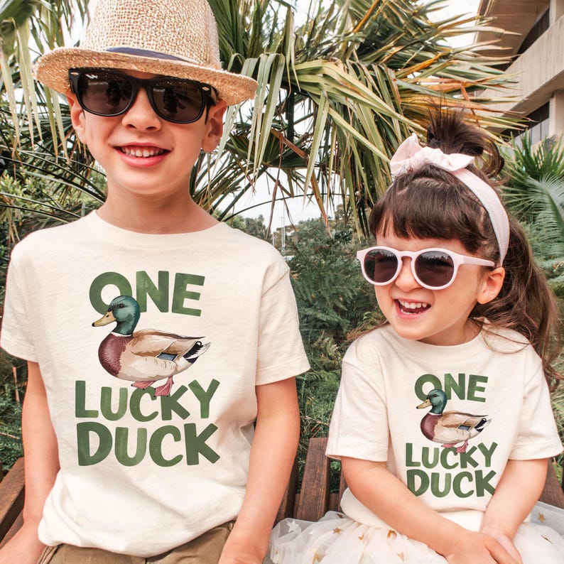 Puede incluir: Dos ni&ntilde;os con camisetas color crema con un gr&aacute;fico de pato y el texto "ONE LUCKY DUCK". El ni&ntilde;o lleva un sombrero de paja y gafas de sol, mientras que la ni&ntilde;a lleva una diadema rosa y gafas de sol. Est&aacute;n sonriendo.