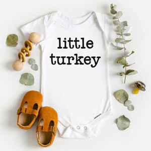 May include: White baby bodysuit with the text "little turkey" in black. The image includes brown baby shoes, a wooden rattle, and sprigs of greenery. A yellow pacifier is also visible.