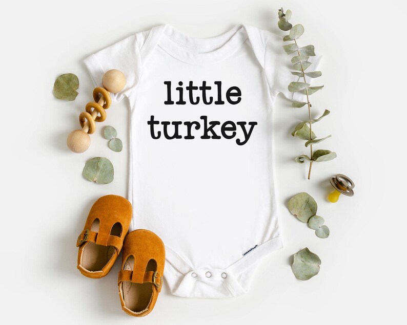 May include: White baby bodysuit with the text "little turkey" in black. The image includes brown baby shoes, a wooden rattle, and sprigs of greenery. A yellow pacifier is also visible.