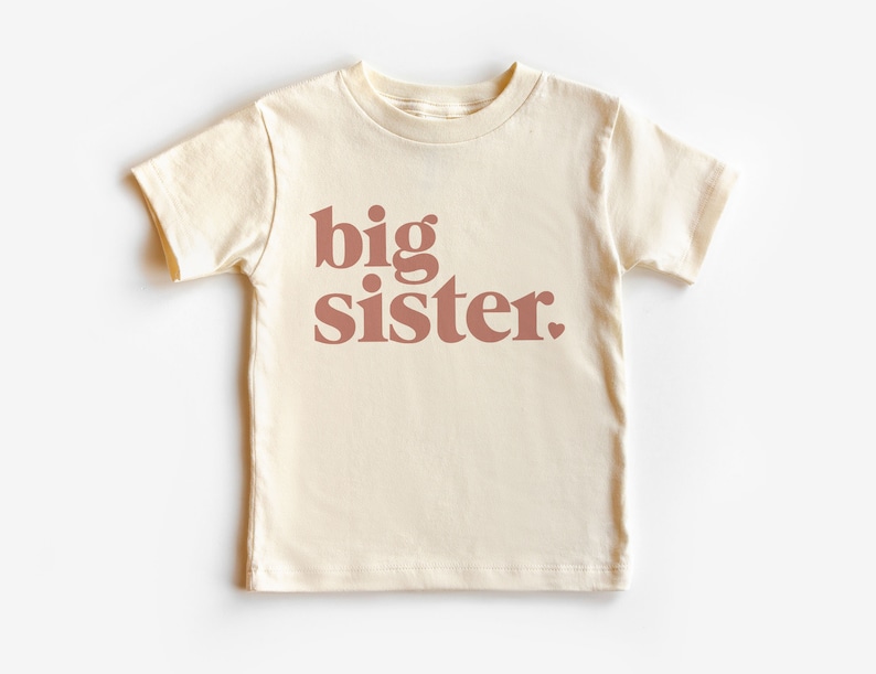 May include: A cream colored short-sleeved t-shirt with the words "big sister" printed in a bold, pink font.