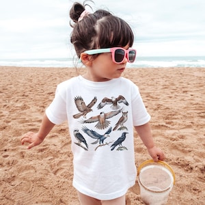 K&ouml;nnte beinhalten: Ein Kind am Sandstrand tr&auml;gt ein wei&szlig;es T-Shirt mit Illustrationen von Raubv&ouml;geln. Das Kind tr&auml;gt eine rosa Sonnenbrille mit mintgr&uuml;nen B&uuml;geln und h&auml;lt einen kleinen gelben Eimer.