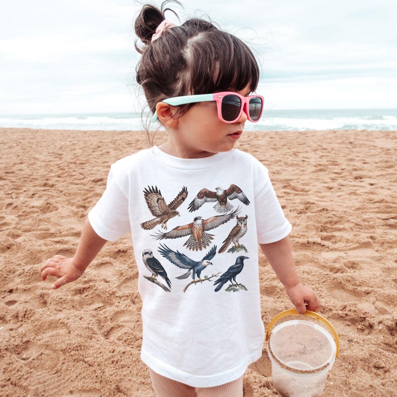 K&ouml;nnte beinhalten: Ein Kind am Sandstrand tr&auml;gt ein wei&szlig;es T-Shirt mit Illustrationen von Raubv&ouml;geln. Das Kind tr&auml;gt eine rosa Sonnenbrille mit mintgr&uuml;nen B&uuml;geln und h&auml;lt einen kleinen gelben Eimer.