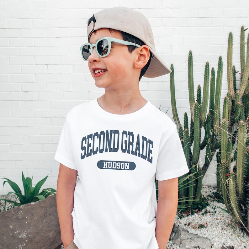 May include: A white t-shirt with the text "SECOND GRADE HUDSON" in navy blue. The image shows a young person wearing the shirt, a beige baseball cap, and mint green sunglasses. The background includes a white brick wall and a cactus.