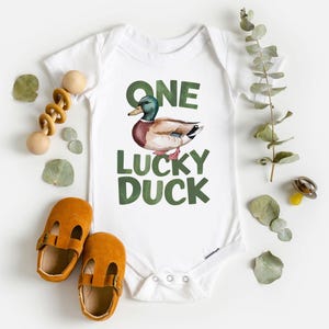 Puede incluir: Body de beb&eacute; blanco con una ilustraci&oacute;n de pato y el texto "ONE LUCKY DUCK" en verde. La imagen incluye zapatos de beb&eacute; naranjas, un juguete de madera y hojas decorativas.
