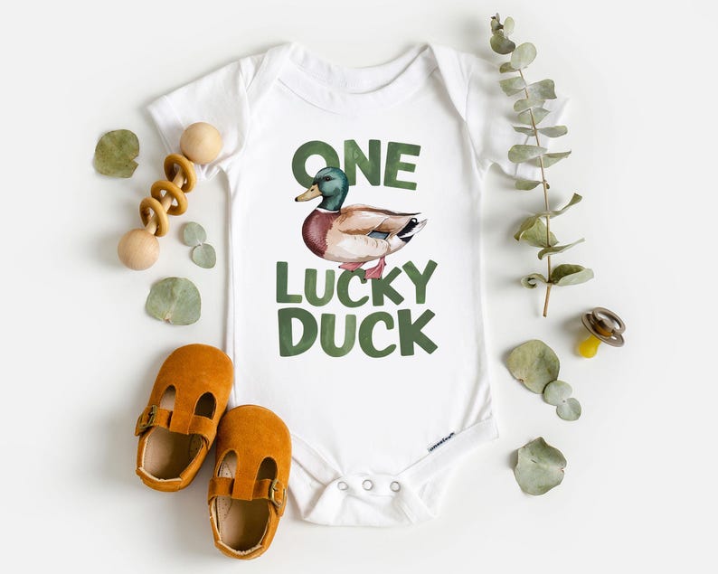 Puede incluir: Body de beb&eacute; blanco con una ilustraci&oacute;n de pato y el texto "ONE LUCKY DUCK" en verde. La imagen incluye zapatos de beb&eacute; naranjas, un juguete de madera y hojas decorativas.