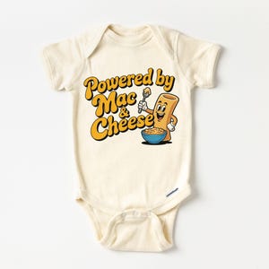K&ouml;nnte beinhalten: Cremefarbener Baby-Body mit dem Text "Powered by Mac & Cheese" in Gelb und Orange. Eine Cartoon-Makkaroni-Figur h&auml;lt einen L&ouml;ffel &uuml;ber einer Sch&uuml;ssel Makkaroni mit K&auml;se. Das Design ist mittig auf der Vorderseite platziert.