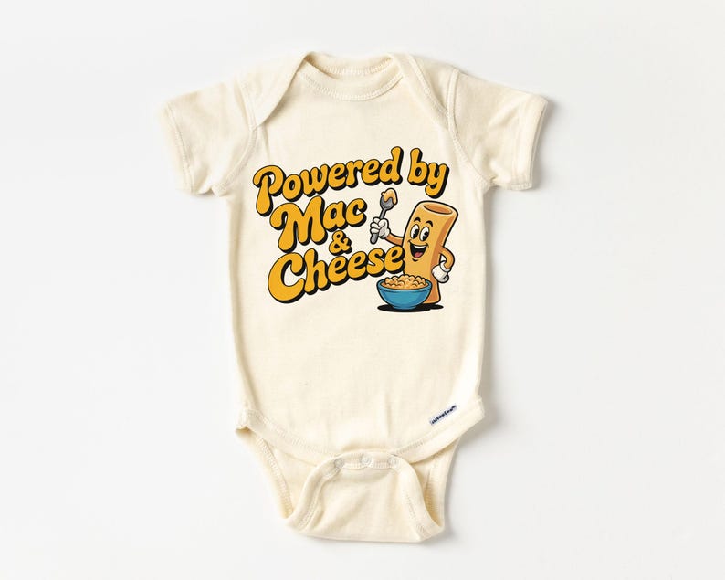 K&ouml;nnte beinhalten: Cremefarbener Baby-Body mit dem Text "Powered by Mac & Cheese" in Gelb und Orange. Eine Cartoon-Makkaroni-Figur h&auml;lt einen L&ouml;ffel &uuml;ber einer Sch&uuml;ssel Makkaroni mit K&auml;se. Das Design ist mittig auf der Vorderseite platziert.