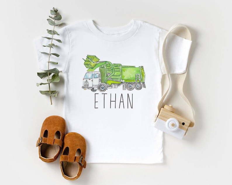 May include: White t-shirt with a green and white watercolor illustration of a garbage truck. The text "ETHAN" is printed below the truck.