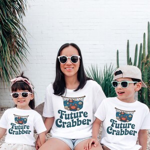 May include: Three people wearing white t-shirts with a "Future Crabber" design. The design features a crab, seashells, and a fishing net. The people are wearing sunglasses and smiling. The background is a white brick wall with plants.
