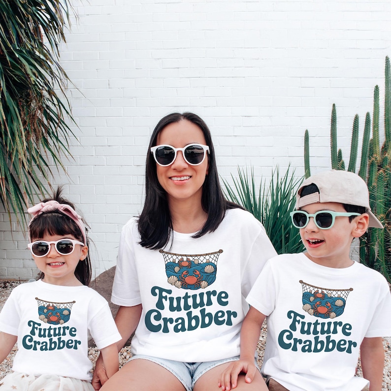 May include: Three people wearing white t-shirts with a "Future Crabber" design. The design features a crab, seashells, and a fishing net. The people are wearing sunglasses and smiling. The background is a white brick wall with plants.