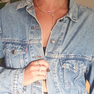 May include: A light blue denim jacket with a classic design, featuring button closures and front pockets. The model is wearing large gold hoop earrings, sunglasses, and a layered necklace. The look is completed with rings and red lipstick.