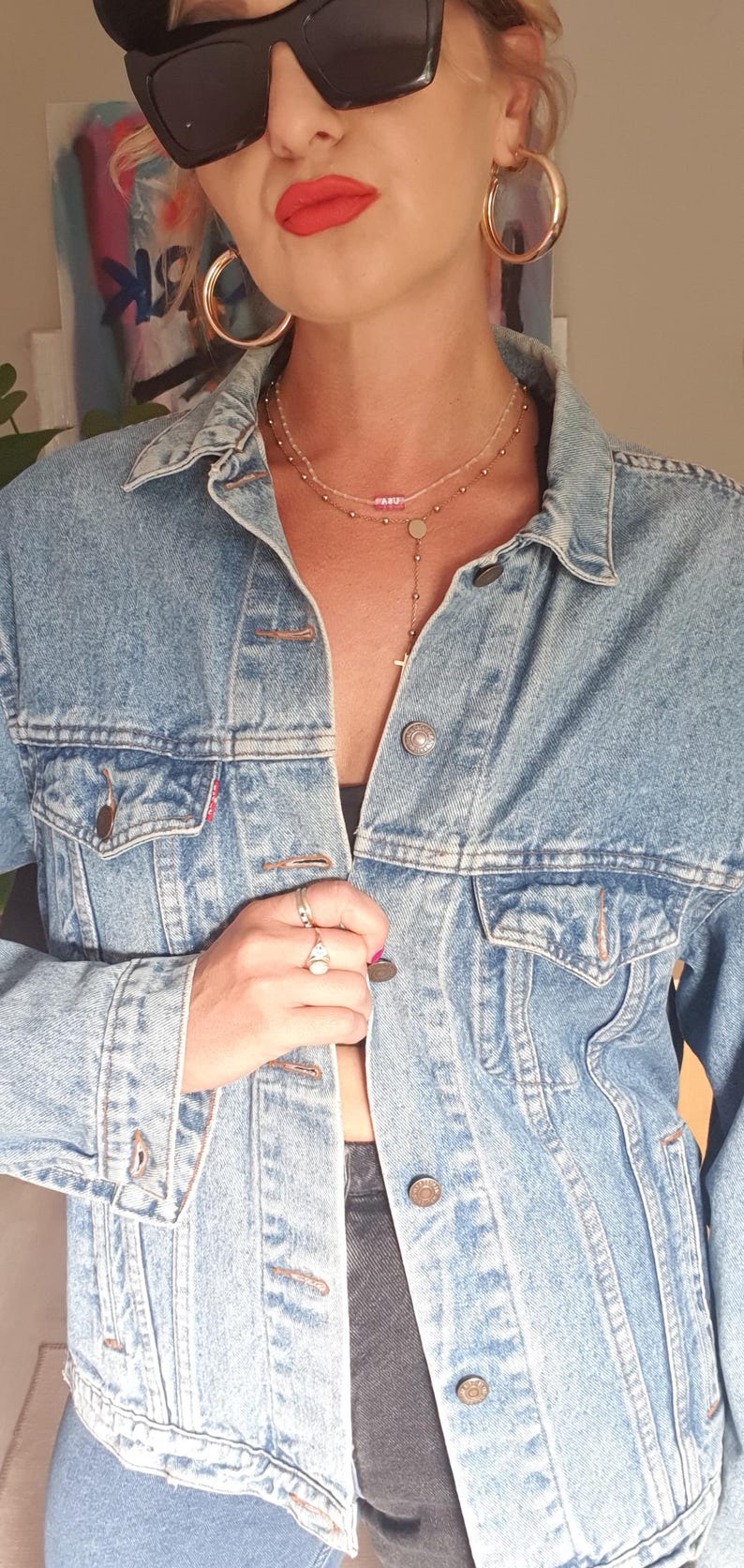 May include: A light blue denim jacket with a classic design, featuring button closures and front pockets. The model is wearing large gold hoop earrings, sunglasses, and a layered necklace. The look is completed with rings and red lipstick.