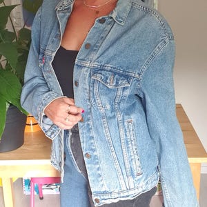 May include: Light blue denim jacket with button closures and chest pockets. The jacket is paired with two-tone jeans, a black cap, and sunglasses. The outfit is completed with leopard print ankle boots and gold hoop earrings.