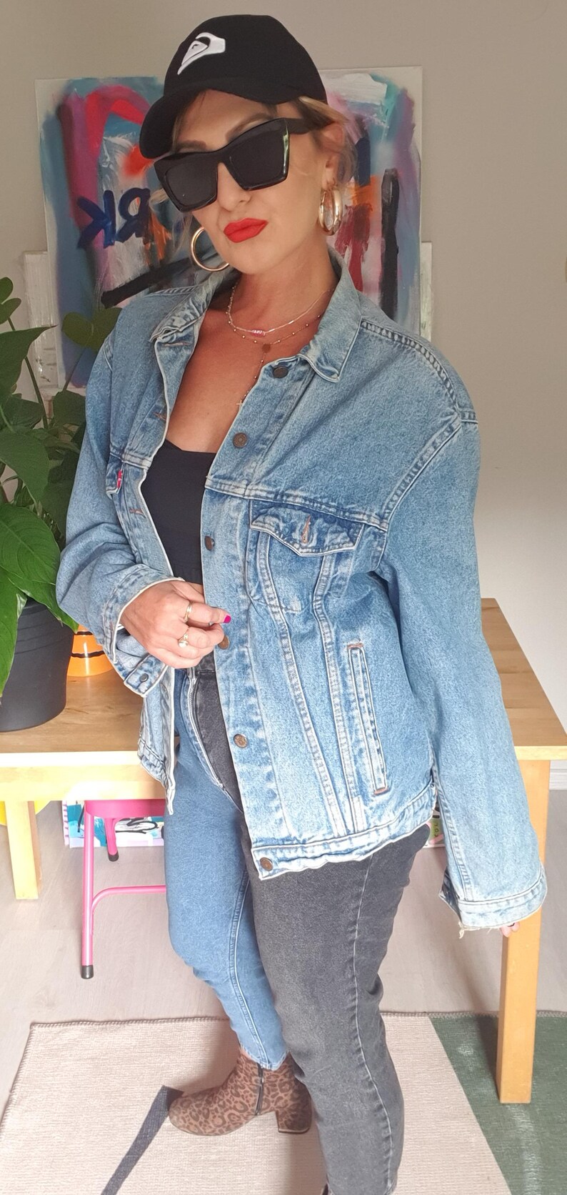 May include: Light blue denim jacket with button closures and chest pockets. The jacket is paired with two-tone jeans, a black cap, and sunglasses. The outfit is completed with leopard print ankle boots and gold hoop earrings.