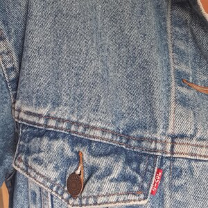 May include: Light blue denim jacket with a button-down front, chest pocket, and a small red tag. The jacket has a classic design with visible stitching and a button closure. The jacket is made of a durable denim material.