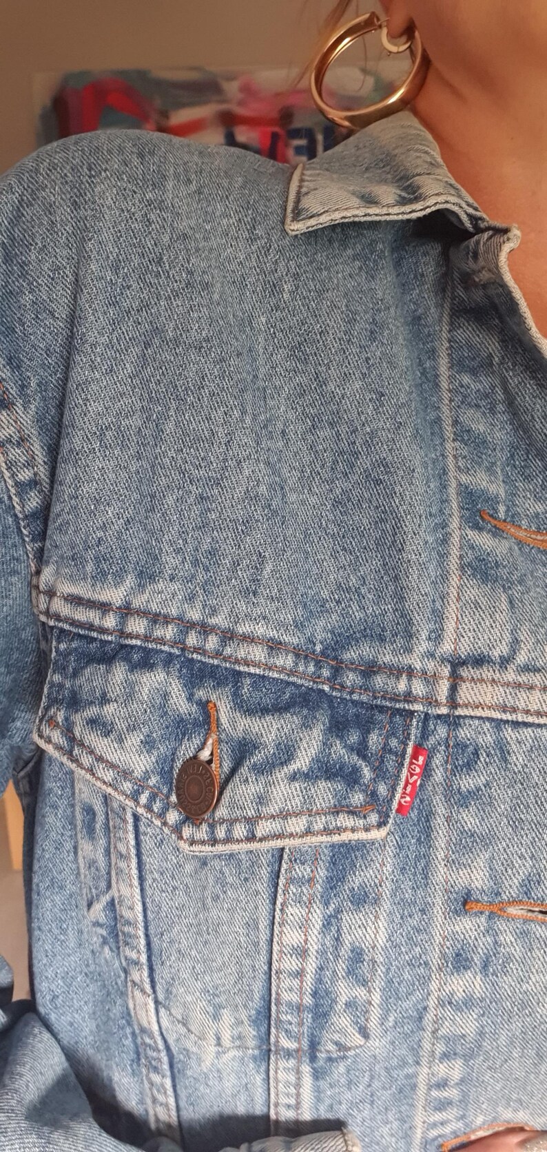 May include: Light blue denim jacket with a button-down front, chest pocket, and a small red tag. The jacket has a classic design with visible stitching and a button closure. The jacket is made of a durable denim material.