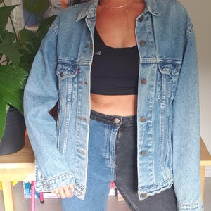 May include: A light-wash denim jacket with a classic collar and button closure. The model is wearing a black crop top, two-tone jeans, a black cap, and sunglasses. The look is completed with gold hoop earrings and a necklace.