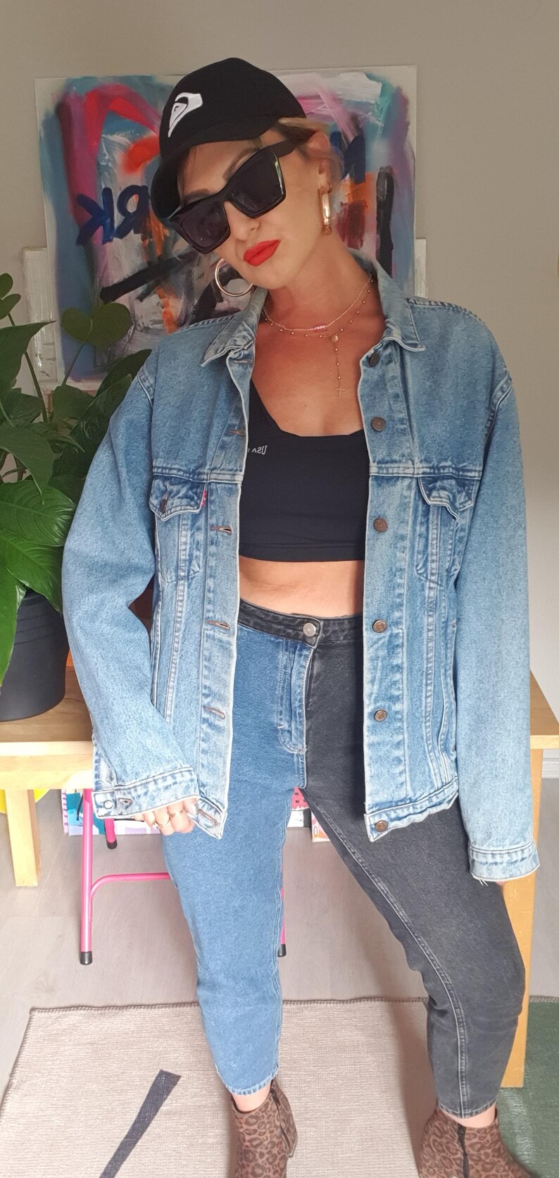 May include: A light-wash denim jacket with a classic collar and button closure. The model is wearing a black crop top, two-tone jeans, a black cap, and sunglasses. The look is completed with gold hoop earrings and a necklace.