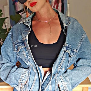 May include: A light blue denim jacket with a black sports bra. The bra has white text. The person is wearing a black cap, sunglasses, and gold hoop earrings. A silver necklace with a cross pendant is visible.