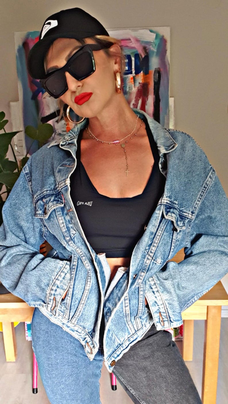 May include: A light blue denim jacket with a black sports bra. The bra has white text. The person is wearing a black cap, sunglasses, and gold hoop earrings. A silver necklace with a cross pendant is visible.