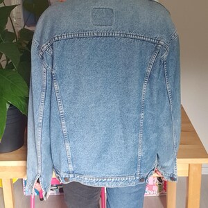 May include: Light blue denim jacket with a classic design. The jacket has a button-up front, a collar, and pockets. The model is wearing a black baseball cap and gold hoop earrings. The jacket is paired with jeans.