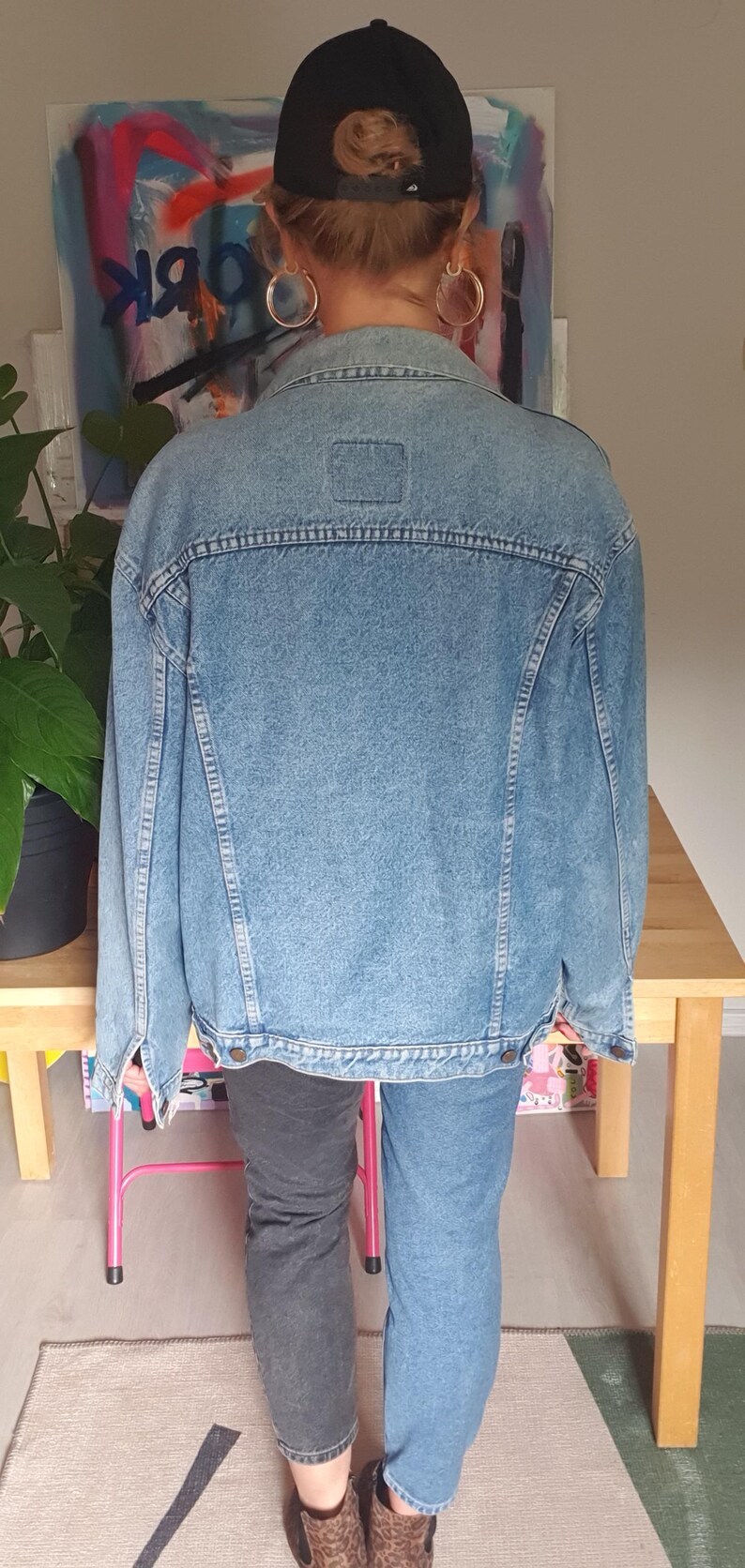 May include: Light blue denim jacket with a classic design. The jacket has a button-up front, a collar, and pockets. The model is wearing a black baseball cap and gold hoop earrings. The jacket is paired with jeans.