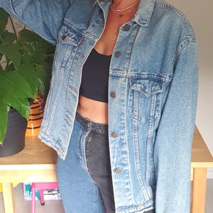 May include: A light blue denim jacket with a button-up front. The person is wearing a black cap, sunglasses, and two-tone jeans. The outfit is completed with leopard print boots and gold hoop earrings.
