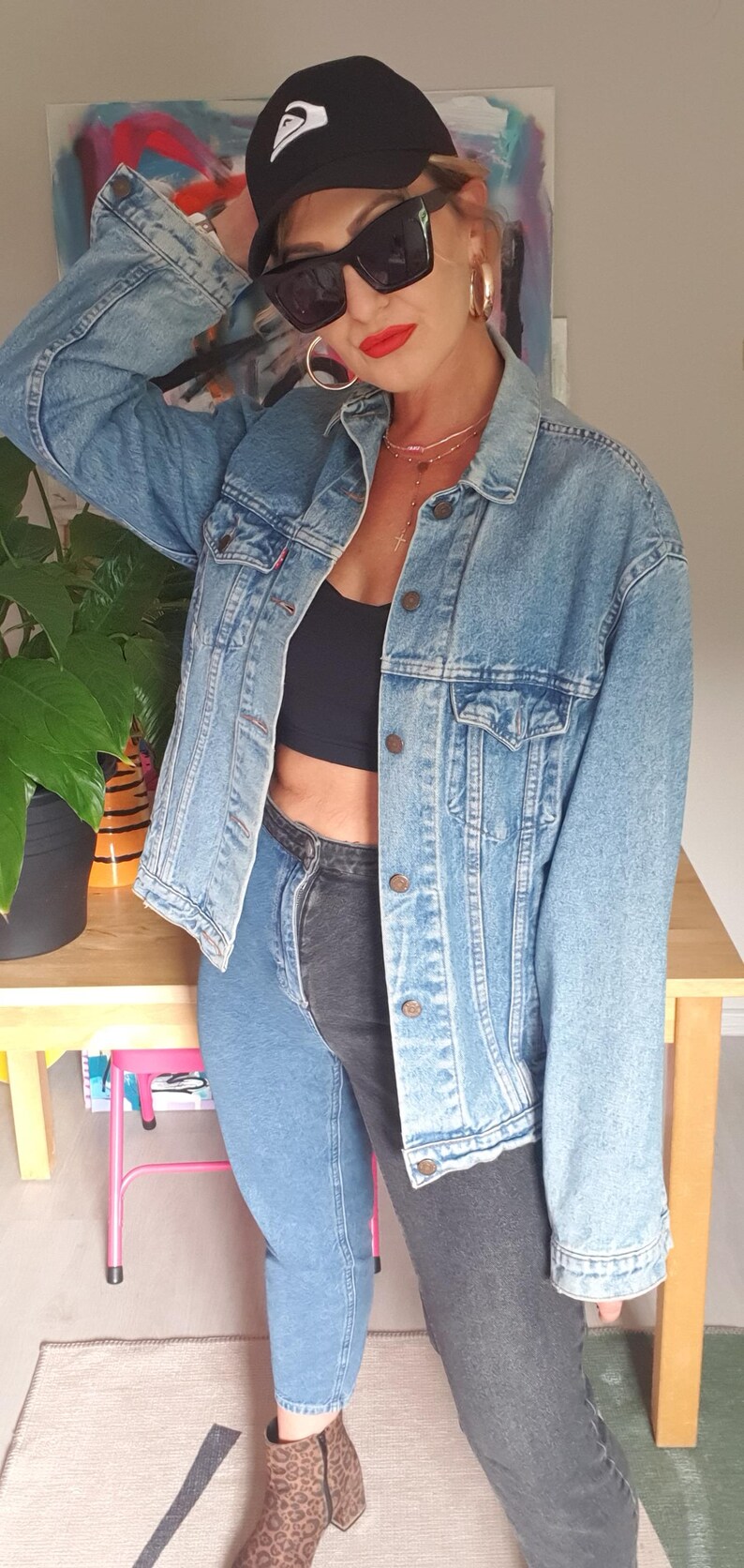 May include: A light blue denim jacket with a button-up front. The person is wearing a black cap, sunglasses, and two-tone jeans. The outfit is completed with leopard print boots and gold hoop earrings.