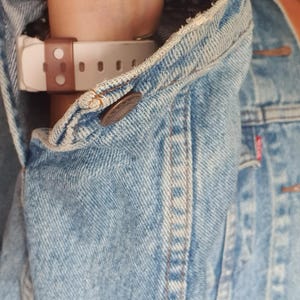 May include: Light blue denim jacket with a buttoned cuff. A white and brown watch is visible under the jacket sleeve. The jacket has a classic design with visible stitching and a button closure.
