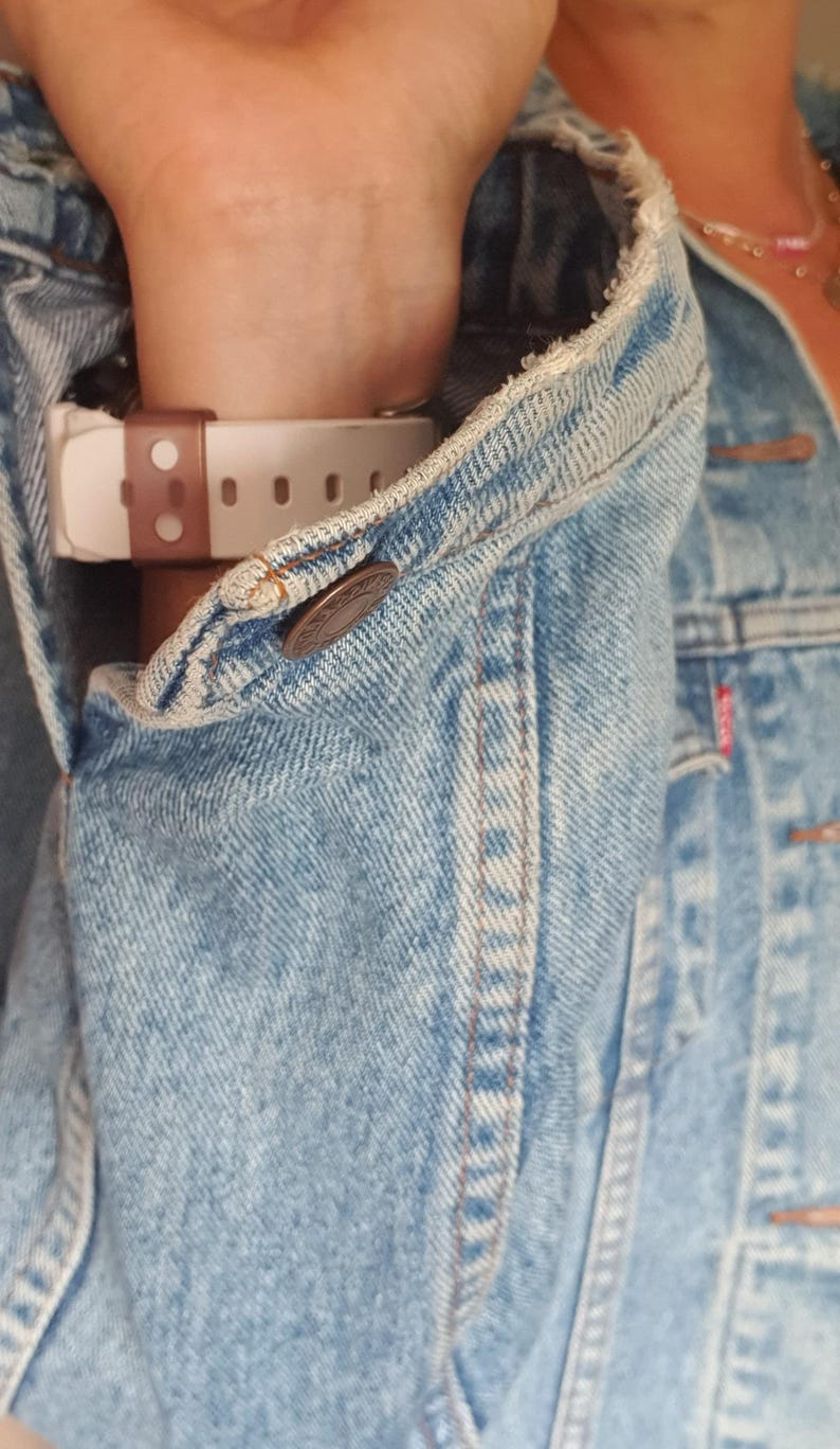 May include: Light blue denim jacket with a buttoned cuff. A white and brown watch is visible under the jacket sleeve. The jacket has a classic design with visible stitching and a button closure.