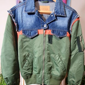 Könnte beinhalten: Grüne Bomberjacke mit blauem Jeanskragen und orangefarbenen Akzenten. Die Jacke hat einen Reißverschluss, zwei Vordertaschen und eine kleine Tasche am Ärmel. Das Etikett am Kragen lautet "SOYA".