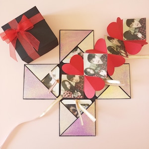 Può includere: Una scatola nera con un nastro rosso e una scatola esplosiva a forma di cuore con foto e cuori rossi. La scatola esplosiva è aperta e le foto sono visibili.