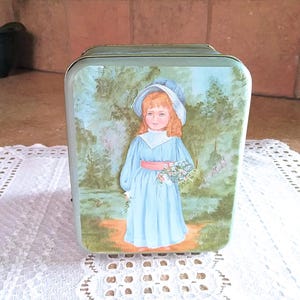 May include: A vintage tin box with a painted image of a young girl in a blue dress and white hat holding a bouquet of flowers. The background is a green and blue landscape.