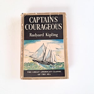 May include: A hardcover book titled Captains Courageous by Rudyard Kipling. The cover features a black background with white text, a central illustration of a sailing ship on blue and white waves, and the text The Great American Classic of the Sea.