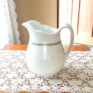 May include: A white ceramic pitcher with a curved handle and a spout. The pitcher has a black and silver band around the middle. The pitcher is sitting on a wooden table covered with a white lace doily.