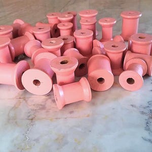 May include: A collection of numerous small, cylindrical wooden spools painted in a solid light pink color. Each spool features a central hole running through its length and flared ends, designed for winding thread or cordage.