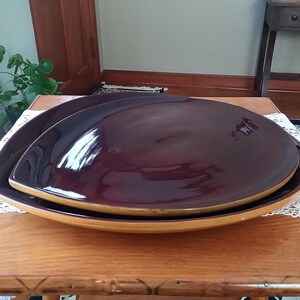May include: Two dark purple ceramic serving dishes, one nested inside the other. The larger dish has a rounded, leaf-shaped rim and a glossy finish. The smaller dish has a similar shape but is slightly smaller.