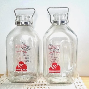 May include: Two clear glass milk bottles with metal handles and lids. Each bottle has a red and black graphic with the words "Equity Top of Ohio" and "Grade A Milk". The bottles are displayed on a white lace doily.