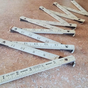 May include: Several vintage folding rulers are scattered on a textured surface. The rulers are white with black markings and numbers, including measurements in feet and inches. Some rulers are partially open, displaying the measurement scales.