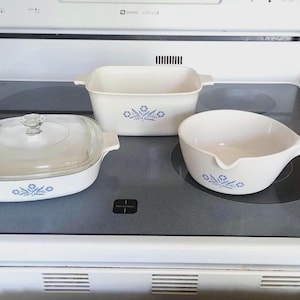 May include: Three white vintage CorningWare dishes with blue floral designs. Includes a square casserole dish with a glass lid, a rectangular baking dish, and a saucepan with a handle.