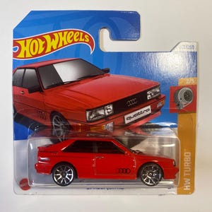 Puede incluir: Coche de juguete rojo Audi Quattro de fundición a presión Hot Wheels con ruedas negras y los logotipos "quattro" y "Audi". El coche está en un blíster de plástico con fondo azul y el logotipo "Hot Wheels".