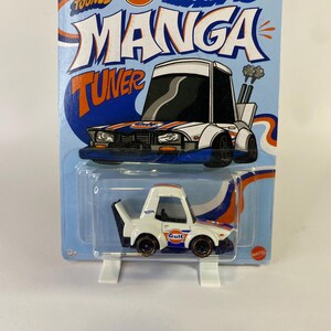 Puede incluir: Un coche de juguete Hot Wheels Manga Tuner blanco y azul con el logotipo de Gulf. El coche está en un paquete azul y blanco con las palabras "Manga Tuner" en letras grandes. El coche tiene detalles en naranja y azul.