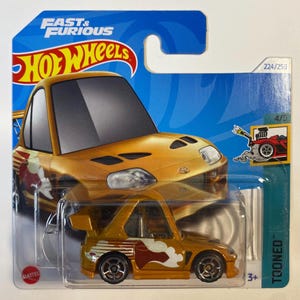 May include: A gold Hot Wheels die-cast toy car with red and white flames on the side. The car is a cartoon version of a Toyota Supra. The packaging shows the car in a more realistic style. The packaging also includes the words "Fast & Furious" and "Toon'd".