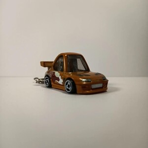 Può includere: Una piccola auto giocattolo arancione con uno spoiler posteriore e una grafica bianca e rossa sul lato. L'auto ha ruote nere e un portachiavi. L'auto è una replica in miniatura di un'auto sportiva.