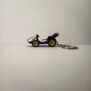 Può includere: Un portachiavi a forma di auto da corsa in miniatura. L'auto è principalmente bianca, blu e rossa, con ruote gialle. È dotato di un cannone nero e un portachiavi argentato. Il portachiavi è progettato per assomigliare a un kart.