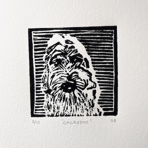 May include: A black and white linocut print depicting a Cockapoo dog's head, set within a black border. The dog's fur is rendered with detailed linework. The print is numbered 2/10, with the word 'cockapoo' and artist's initials.