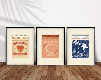 May include: Three framed prints of matchbox labels. The first is red and white with a heart and the text "FIREHEART YOU DO NOT YIELD". The second is pink and white with a crescent moon and the text "LIGHT IT UP PRESCERT CITY SAFETY MATCHES". The third is blue and white with a star and the text "STARFALL SMILE AGAIN".